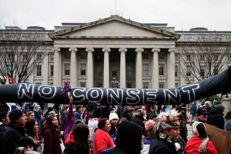 A large protest march with dozens of people holding up a mock oil pipeline that reads 'No Consent'. The U.S. Treasury building is in the background.